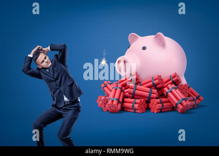 Businessman se protéger avec les mains et pink piggy bank sur rouge bâtons de dynamite avec fusible sur fond bleu Banque D'Images