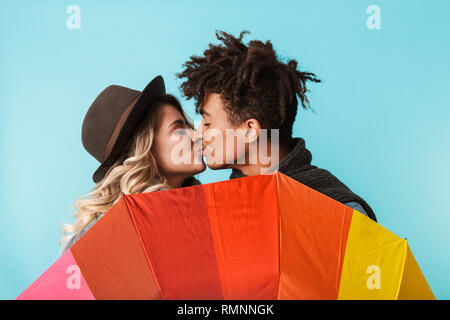 Heureux couple multiracial portant des vêtements d'hiver debout, tenant un parapluie isolé sur fond bleu, les baisers Banque D'Images