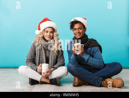 Teenage couple sitting multiraciale heureux isolé sur fond bleu, portant des chapeaux de noël, holding tasses Banque D'Images