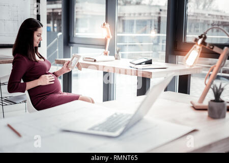 Baby bump. Les jeunes appel dark-haired woman feeling sentimental tout en tenant son bébé bosse dans un salon spacieux Banque D'Images
