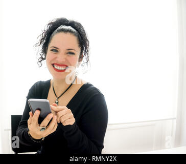 L'âge moyen à la normale caucasian woman jouer avec un smartphone en souriant et regardant la caméra. Lèvres rouge grand sourire, cheveux noirs et yeux bleus très heureux Banque D'Images