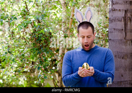 Une surprise de Pâques ! Homme tenant un oeuf de Pâques en or avec des oreilles de lapin sur la tête. Banque D'Images