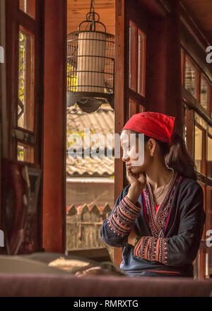 Une femme Dao rouge dans les vêtements brodés minutieusement regards par un fenêtre verticale dans un bâtiment traditionnel à Sapa, Vietnam. Banque D'Images