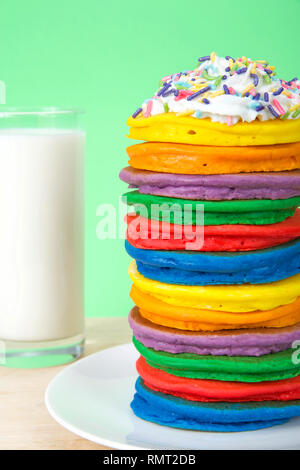 Pile de crêpes colorées lumineuses garnies de crème fouettée et saupoudre de bonbons avec un fond vert et verre de lait. Présentation verticale, Fairy Banque D'Images