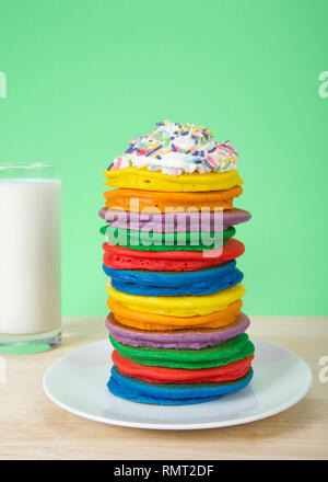 Pile de crêpes colorées lumineuses garnies de crème fouettée et saupoudre de bonbons avec un fond vert et verre de lait. Présentation verticale, Fairy Banque D'Images