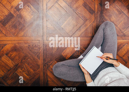 Concept de travail confortable à la maison. Vue de dessus woman sitting on wooden floor écrit dans le bloc-notes, copier l'espace. Banque D'Images