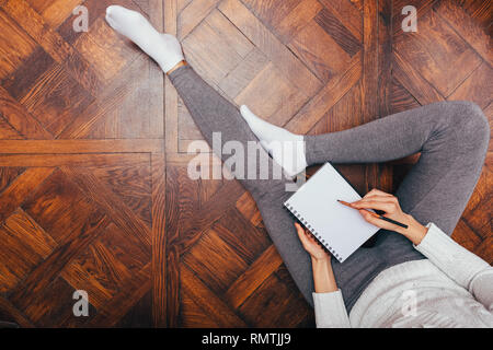 Concept de travail confortable à la maison. Vue de dessus woman sitting on wooden floor écrit dans le bloc-notes. Banque D'Images