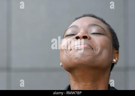 Heureux African American Woman portrait en gros plan, la tête rejetée en arrière dans un sourire. Debout contre un mur gris background with copy space Banque D'Images