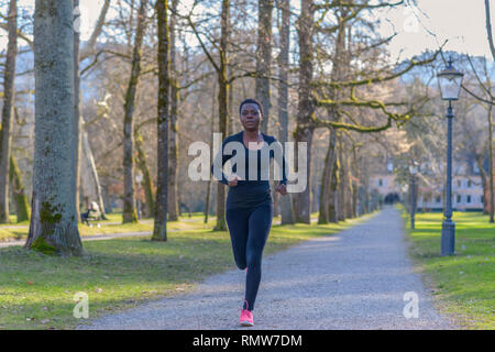 Jeune femme africaine le jogging pendant son entraînement quotidien en marche le long d'une allée bordée d'arbres dans un parc urbain près de l'appareil photo Banque D'Images