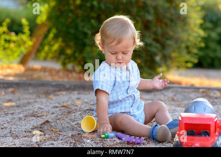 Adorable petit garçon blond bébé joue dehors avec ses jouets en plastique coloré assis sur le sol dans un portrait Banque D'Images