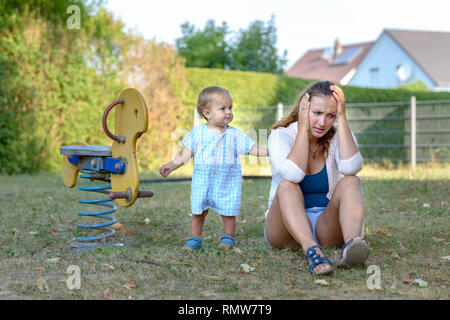 Jeune bébé fils réconfortant sa mère a souligné que elle s'assoit sur le sol dans une aire de jeux avec lui dans un moment de tendresse de l'amour une relation Banque D'Images