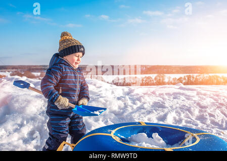 Petit garçon avec une pelle à neige en hiver dans le parc. Heureux qui peut accueillir la neige en tubes, dans des vêtements décontractés. Banque D'Images
