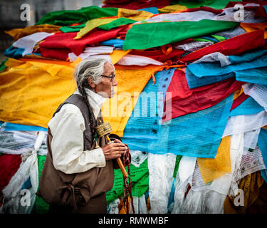 Lhassa, Tibet. 20 juillet 2016. Femme tibétaine spining son terminal mobile à prières passe devant un fond plein de drapeaux de prière aux couleurs vives. Banque D'Images