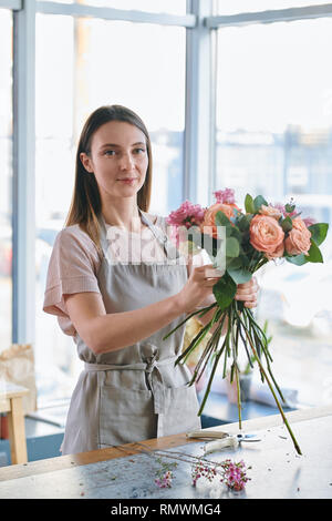 Jolie fille en tablier, debout par table en face de la caméra et vous regarde tout en bouquet de roses Banque D'Images