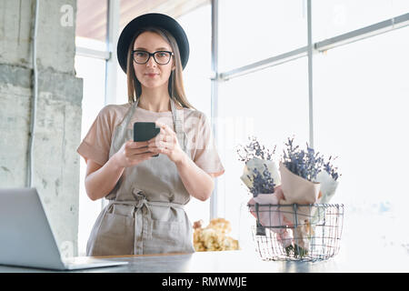 Jeune femme en vêtements de travail, lunettes et chapeau en défilement tout en travaillant dans smartphone studio floral Banque D'Images