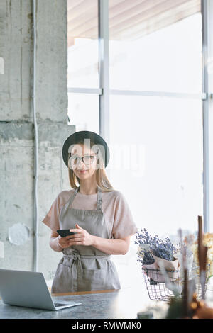 Jeune femme élégante avec chapeau et workwear standing par table pendant le défilement dans le smartphone en studio fleuriste Banque D'Images