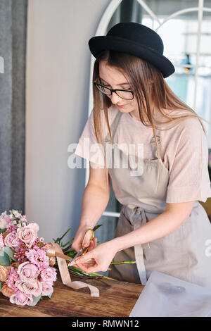 Jeune et Jolie fleuriste beige coupe ruban de soie se termine avec des ciseaux tout en bouquet de fleurs rose pastel en studio Banque D'Images