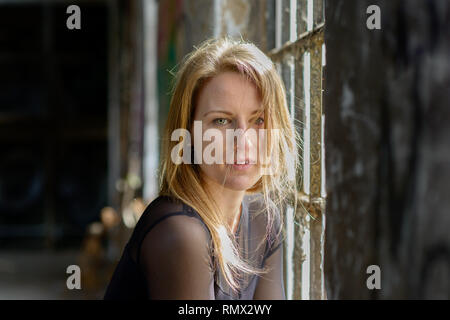 Belle fille blonde avec des yeux verts et des cheveux ébouriffés longue fixant songeur à l'appareil photo avec parted lèvres aux côtés d'une vieille fenêtre à l'intérieur rustique Banque D'Images