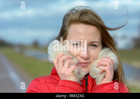 Jolie jeune femme blonde à l'extérieur en hiver se pelotonnant dans un endroit chaud garni de fourrure veste rouge à côté de la piste d'un pays sur le long d'une rivière Banque D'Images