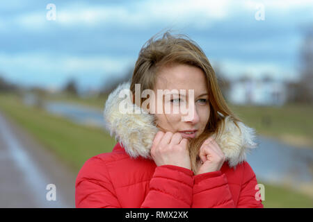 Jolie jeune femme blonde à l'extérieur en hiver se pelotonnant dans un endroit chaud garni de fourrure veste rouge à côté de la piste d'un pays sur le long d'une rivière Banque D'Images