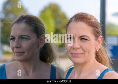 Femme d'âge moyen reflète dans la fenêtre d'un immeuble commercial comme elle se tient avec sa joue au regard de l'avant en verre avec un sourire tranquille Banque D'Images