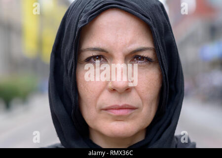 Pensive woman portant un foulard noir fixant intensément la caméra dans un cropped head shot à l'extérieur, sur une rue urbaine Banque D'Images