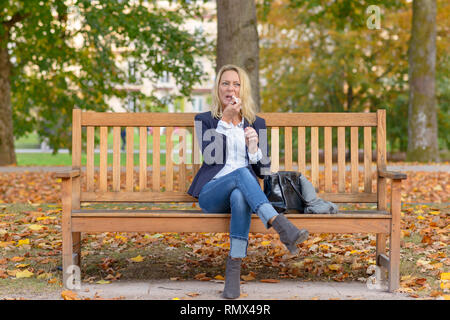 Trendy smiling blonde woman using her lipstick assis sur un banc en bois Banque D'Images