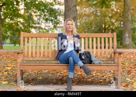 Trendy smiling blonde woman using her lipstick assis sur un banc en bois Banque D'Images