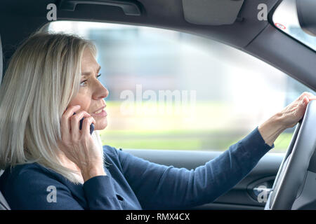 Femme mature chat pilote sur son portable dans la voiture qu'elle conduit à travers la ville dans une vue de côté à travers la vitre du passager Banque D'Images