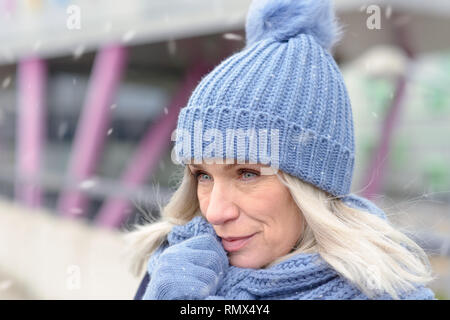Jolie femme blonde dans une tenue à la mode d'hiver bleu se pelotonnant dans une écharpe tenant son les mains gantées dans la chute de neige d'hiver Banque D'Images