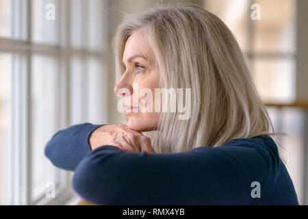 Belle femme blonde en regardant à travers une vitre avec une expression grave posant son menton sur ses mains alors qu'elle s'appuie sur une balustrade de bois Banque D'Images