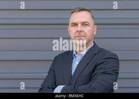 Homme d'âge moyen confiant dans blazer veste avec bras croisés contre un mur gris en vue de côté avec copie espace Banque D'Images