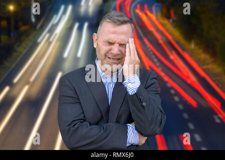 Stressed businessman, ou souffrant d'un mal de tête, debout avec sa main à son temple des grimaces contre la longue exposition light trails de trafic sur un Banque D'Images