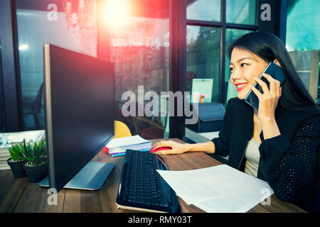 Jeune femme asiatique au visage souriant à pleines dents en conversation sur smart phone et la tenue de la souris d'ordinateur Banque D'Images