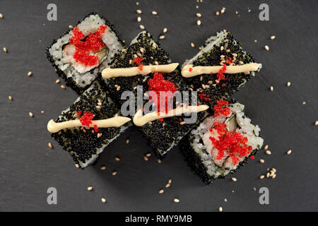 Philadelphia maki sushi au fromage à la crème, le concombre et les poissons. Décorées avec rouleau de mayo, tobiko rouge re et sésame. Vue de dessus, servi sur la plaque en pierre noire. Banque D'Images