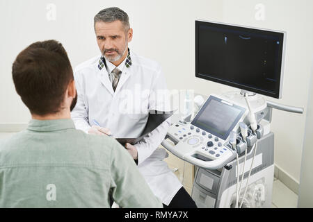 Patient assis dans le cabinet médical avec médecin professionnel et consulting. Thérapeute sérieux portant en robe blanche médicale parler, maintenant titulaire. L'équipement moderne pour l'échographie. Banque D'Images