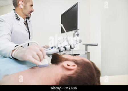 L'ultrasonographie, le diagnostic échographique des ganglions sur le cou. Homme barbu allongé en cabinet médical. Woman in white, blouse et des gants médicaux d'examen faisant avec sonde échographique. Banque D'Images