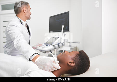 Homme africain couché dans le cabinet médical. L'ultrasonographie, le diagnostic échographique des ganglions sur le cou. Woman in white, blouse et des gants médicaux d'examen faisant avec sonde échographique. Banque D'Images