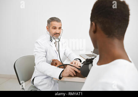 Médecin à l'aide d'un sphygmomanomètre la mesure de la pression artérielle du patient. African man sitting on consultation, tenant la main sur le tableau. Docteur en médecine en robe blanche. Banque D'Images
