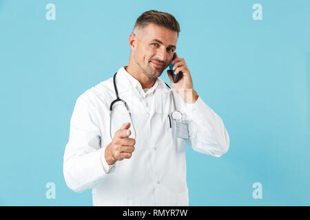 Photo de médecin positive portant blouse blanche et stéthoscope talking on cell phone isolés sur fond bleu Banque D'Images