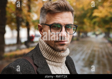 Photo de l'homme adulte 30s portant des lunettes à la caméra à l'extérieur tout en marchant à travers le parc en automne Banque D'Images