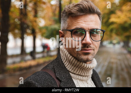 Photo de l'homme masculin 30s portant des lunettes à la caméra à l'extérieur tout en marchant à travers le parc en automne Banque D'Images