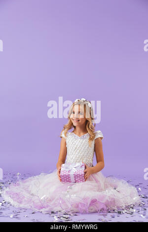 Jolie petite fille vêtue de robes de princesse assis sur fond violet isolés, holding present fort Banque D'Images