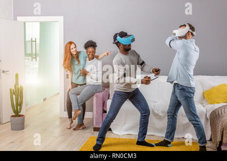 Diversité multiethnique friends enjoying VR technology, deux gars portant casque vr et à jouer tout en African and Caucasian girl rousse regarde et Banque D'Images