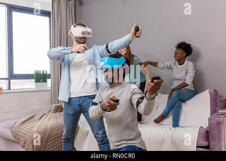 Les étudiants internationaux au repos avec divers lunettes de réalité virtuelle d'une maison, deux jeunes hommes filles interracial divertissant. Banque D'Images