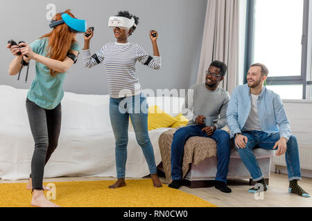 Deux jeunes femmes multiraciales portant des lunettes de réalité virtuelle de danse à la maison pendant que leurs petits amis assis sur le canapé et souriant, profitant de l'action Banque D'Images