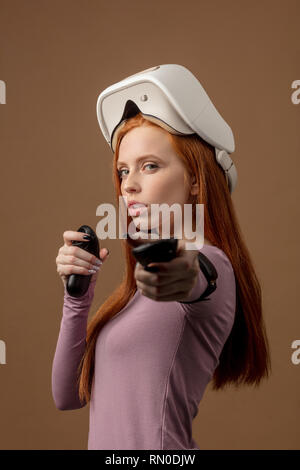 Portrait rousse jeune femme looking at camera with non-active affichage tête montée sur la tête, visant à huis clos tenue joysticks en mains. Virtial Banque D'Images