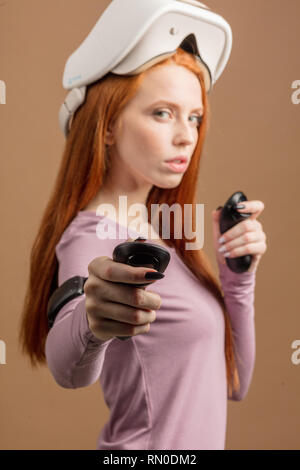 Portrait rousse jeune femme looking at camera with non-active affichage tête montée sur la tête, visant à huis clos tenue joysticks en mains. Virtial Banque D'Images