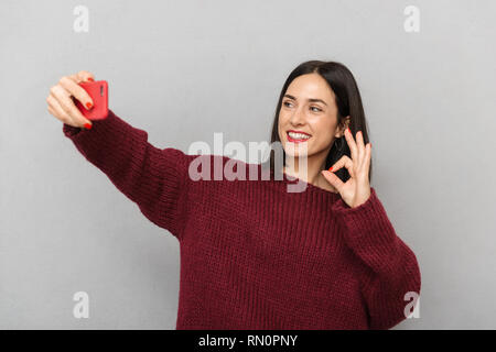Photo de jolie jeune femme vêtue de chandail bourgogne selfies prendre par téléphone mobile au fond isolé mur gris. Banque D'Images