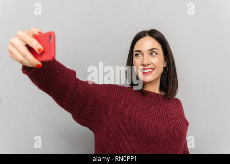 Photo de jolie jeune femme vêtue de chandail bourgogne selfies prendre par téléphone mobile au fond isolé mur gris. Banque D'Images